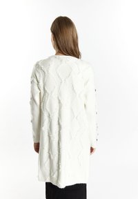 Donna con i capelli castani lisci che indossa un lungo cardigan bianco a maglia testurizzata sopra una gonna nera, vista da dietro su uno sfondo bianco.