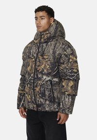 Kurtka puchowa w camo z wysokim kołnierzem, z mieszanką brązowych, zielonych i szarych wzorów liści, kieszeniami na zamek i pikowanym designem.