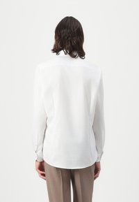 Chemise à manches longues, blanche, en tissu lisse. Caractérisée par un col pointu, des poignets boutonnés et un ourlet arrondi à l'arrière.