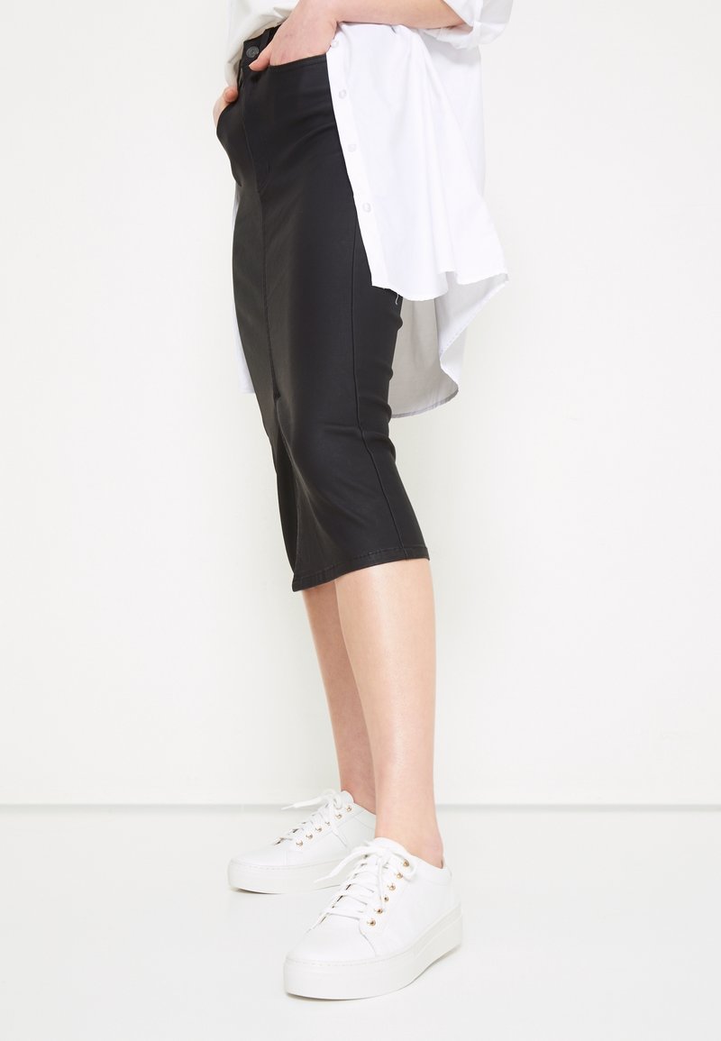 Personne portant un pantalon noir ajusté aux genoux, des baskets blanches basses et une longue chemise blanche avec les mains dans les poches.