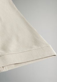 Manga de tela ribeteada en beige claro, con corte recto y puño doblado. Textura suave y ligero brillo con un diseño limpio y minimalista.