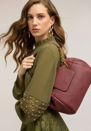 Kvinde med bølget brunt hår iført guldbelagte øreringe og en grøn bluse med blonder på ærmerne, som bærer en tekstureret vinrød håndtaske med guldkæde-stropper.