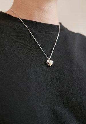 Silver heart pendant necklace on a silver chain, worn over a black t-shirt. Pendant is textured with curved lines and a reflective surface.