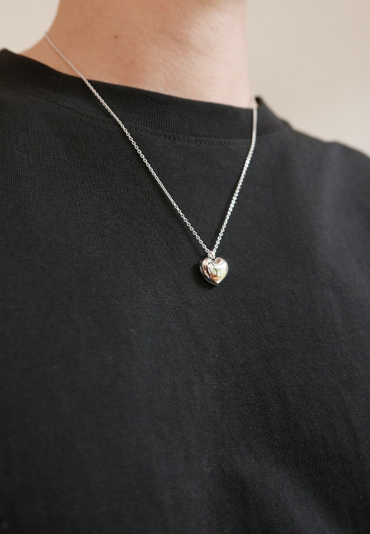 Pandantiv argintiu în formă de inimă pe un lanț argintiu, purtat peste un tricou negru. Pandantivul are o textură cu linii curbe și o suprafațăreflectorizantă.