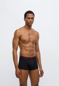 Boxers pretos com um logótipo vermelho "HUGO" na cintura. O tecido parece suave e o ajuste é justo ao corpo.