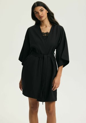 Młoda kobieta z długimi ciemnymi włosami, ubraną w czarny kimono z paskiem, z koronkowym zdobieniem przy dekolcie, stojąca na jasnym, jednolitym tle.