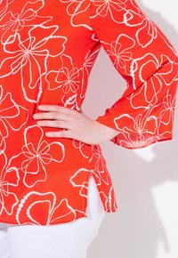 Blouse rouge avec des motifs floraux blancs, manches longues transparentes, fentes sur les côtés au niveau de l'ourlet et une texture lisse. Positionnée avec une main sur la taille.
