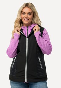 Gilet nero con accenti trapuntati e zip bianca, indossato sopra una felpa leggera color viola chiaro con una consistenza particolare. Jeans in denim chiaro.