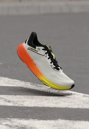 Une chaussure de course unique avec une tige blanche, des lacets noirs et une semelle dégradée orange-jaune, capturée en plein air au-dessus d'une surface asphaltée.