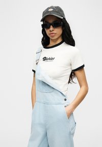 Νεαρή γυναίκα με γκρι καπέλο, μαύρα γυαλιά ηλίου, λευκό t-shirt με μαύρο γιακά και λογότυπο, και ανοιχτό μπλε τζιν σαλοπέτα με ένα λουρί χαλαρό.