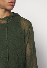 Top con cappuccio verde realizzato in tessuto a maglia trasparente e texturizzato, con una vestibilità ampia. Presenta un cordino e elementi di design a strati visibili.