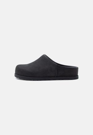 Sabot slip-on negru cu vârf închis și spate deschis, având talpă plată și un logo subtil al mărcii pe lateral.