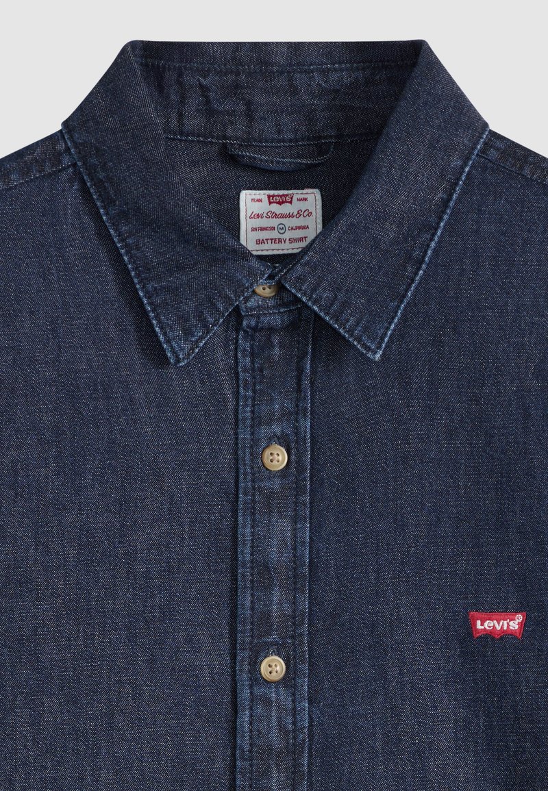 Chemise en denim bleu foncé avec col pointu, boutons beige et petit écusson rouge avec le logo Levi's sur la poche poitrine.