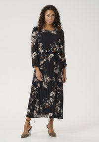 Garais melnais kleitas ar krēmkrāsas un brūnu ziedu rakstu, plisēts dizains, apaļš kakla izgriezums un garas piedurknes, kombinēts ar bēšām augstpapēžu kurpēm.