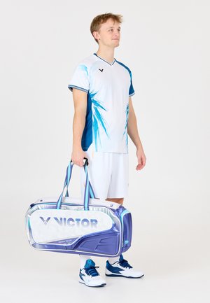 Ung mand i sportstøj, der holder en stor VICTOR sportstaske, stående foran en ensfarvet hvid baggrund.