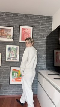 Svetlo siva dukserica i odgovarajuće trenirke, ležeran kroj, teksturirani materijal, elastičan pojas, naslonjena je na sivu zidnu opeku sa šarenim umetničkim radovima.