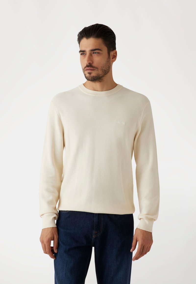 Pull à manches longues beige clair en tricot texturé avec un col rond et des poignets côtelés. Détail de logo subtil sur la poitrine.