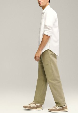 Chemise blanche à manches longues, pantalon kaki avec poches latérales et baskets marron avec des accents crème. Le modèle se tient de profil contre un fond neutre.