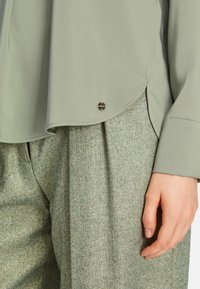 Hellgrüne Bluse aus glattem Stoff mit abgerundetem Saum, ausgestattet mit einem markierten Metallelement. Kombiniert mit strukturierten grünen Hosen.
