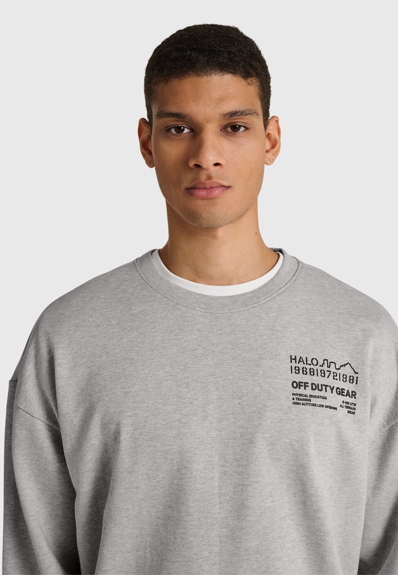 Junger Mann mit kurzen Haaren, der ein hellgraues Sweatshirt mit schwarzem Text "HALO OFF DUTY GEAR" auf der linken Brust vor einem einfarbigen Hintergrund trägt.