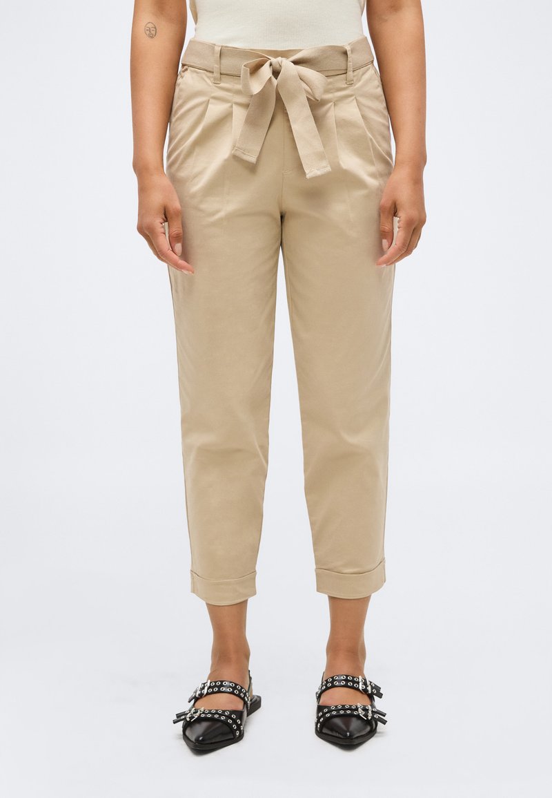 Μπεζ ψηλόμεσοι cropped παντελόνες με δέσιμο στη μέση, φορεμένες με μαύρα μυτερά φλατ παπούτσια με διπλή αγκράφα και λουράκια με οπές.