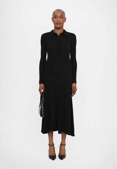 Garās melnas kleitas, kas izgatavota no ribota auduma, priekšpusē ar pogām, garām piedurknēm un izsēdējušo apakšmalu. Kombinēta ar teksturētu zaļu rokassomu.