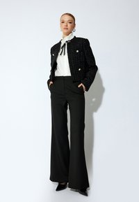 Giacca cropped in tweed nera con bottoni argentati, camicia bianca a balze e pantaloni neri a gamba larga. Décolleté neri a punta completano il look.