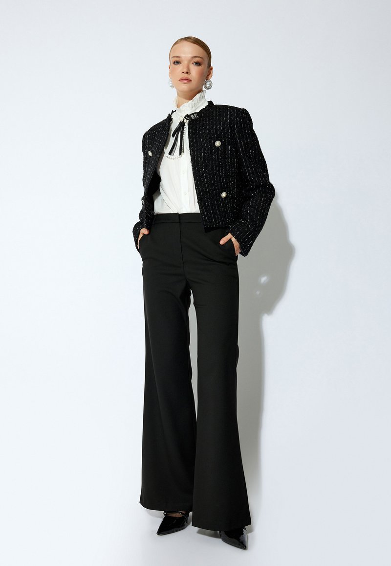 Giacca cropped in tweed nera con bottoni argentati, camicia bianca a balze e pantaloni neri a gamba larga. Décolleté neri a punta completano il look.