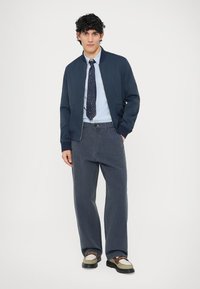 Tengerészkék bomberdzseki, világoskék ing, mintás nyakkendő, bőszárú szürke nadrág és kéttónusú cipők fekete talppal. Simább anyag és texturált részletek.