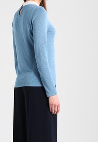 Ljusblå långärmad topp med vit krage och en liten nyckelhålsdetalj på baksidan. Mjuk textur; matchad med mörka byxor.