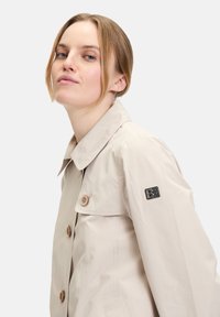 Beiger Trenchcoat mit Kragen, zwei großen Knöpfen und einem Patch mit "BB" am Ärmel. Glatte Textur und taillierte Passform.