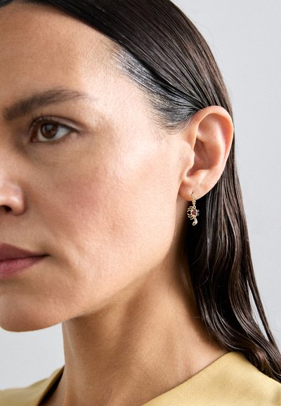 Pendientes en tono dorado con un diseño intrincado que presenta una pequeña piedra púrpura, colgando de un delicado aro. El cabello está peinado hacia atrás.