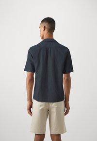 Cămașă cu mânecă scurtă de culoare gri închis, dintr-un material texturat, cu guler clasic și detalii la spate, asortată cu pantaloni scurți de un bej deschis.