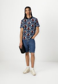 Kortermet polo skjorte med et blått geometrisk mønster og røde detaljer, kombinert med denimshorts og lysfargede joggesko. Holder en svart veske.