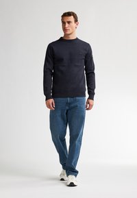 Donkerblauwe trui met ronde hals gemaakt van gestructureerde stof, met een klein logopatchje op de borst. Gecombineerd met losse denim jeans en sneakers.