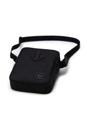 Black fabric shoulder bag with a square shape, textured finish, adjustable strap, and embossed logo patch on the front.