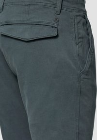 Pantalon en coton vert foncé avec une poche arrière à rabat, tissu texturé et détails de couture subtils.