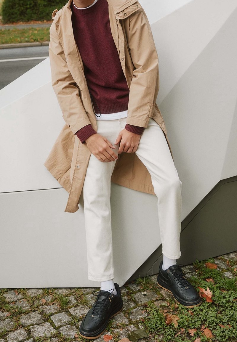 Manteau imperméable beige sur un pull bordeaux, assorti à un pantalon blanc et des baskets noires. Caractéristiques incluent une capuche et des poignets côtelés, sur fond géométrique.