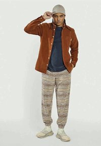 Camisa de corduroy castanha, t-shirt azul-escura, calças de jogging beje com padrão, gorro de malha cinzento e sapatilhas claras. Ajuste simples e descontraído.