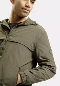 Jasnozielona, lekka kurtka z kapturem, zapinana na zamek, z elastycznymi mankietami i subtelną naszywką z logo na rękawie, charakteryzująca się gładką teksturą materiału.