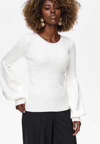 Vrouw met krullend haar, grote oorbellen, een witte geribbelde trui met pofmouwen en zwarte, wijd uitlopende broek tegen een witte achtergrond.