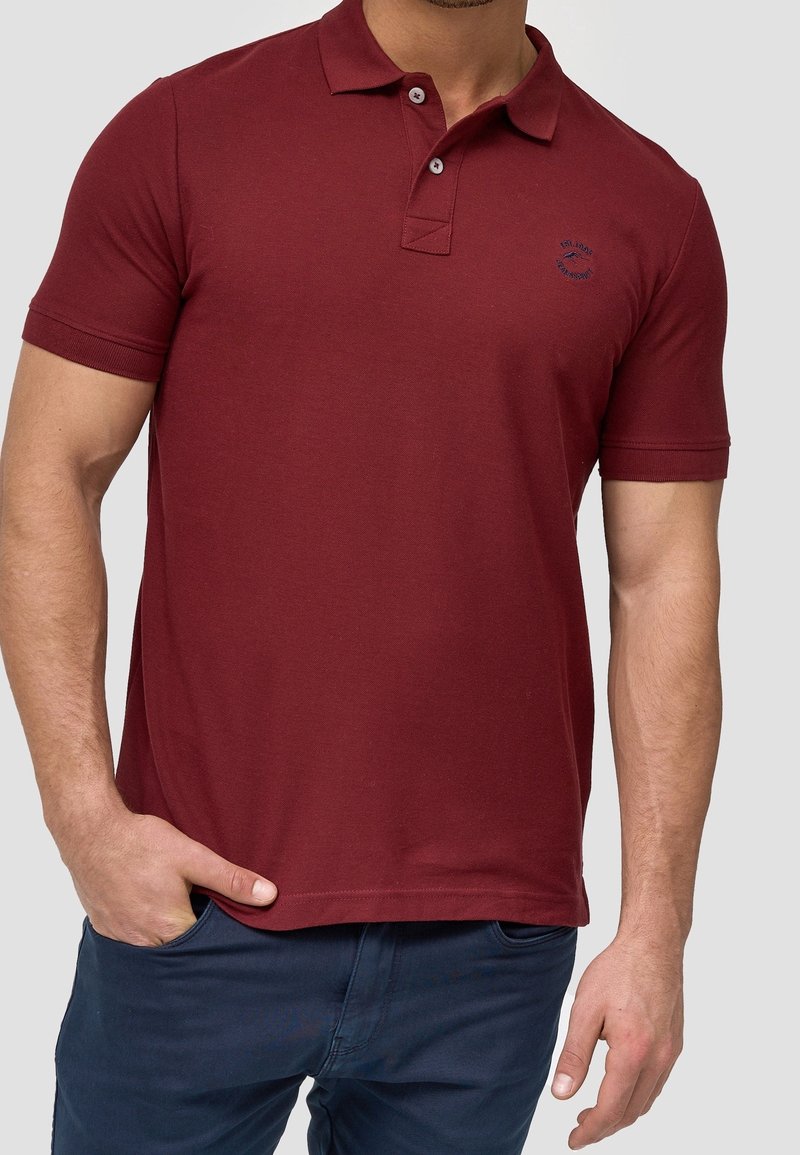 Homme portant un polo bordeaux à manches courtes avec deux boutons et un pantalon bleu foncé, une main dans la poche.