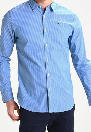 Man wearing a fitted light blue striped long-sleeve button-up shirt and dark pants, standing with one hand slightly raised.