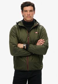 Olivgrön lättviktsjacka med huva, med en röd dragkedja och ett logopatch. Tyget verkar slätt och glänsande, med elastiska ärmslut.