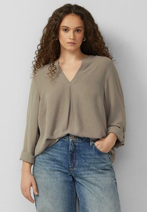 Woman with curly hair wearing a loose beige V-neck blouse with rolled-up sleeves and blue high-waisted jeans, hand in pocket.