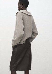 Maglione beige a coste in maglia con un ampio colletto, abbinato a una gonna a matita marrone in pelle, visto da dietro. Tessuto liscio e sagoma aderente.