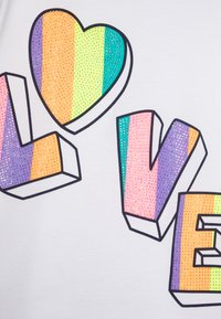 Hvit t-skjorte med regnbuefargede, teksturerte "KJærlighet"-bokstaver, med en hjerteform. Bokstavene har en shiny, rhinesten-lignende finish.