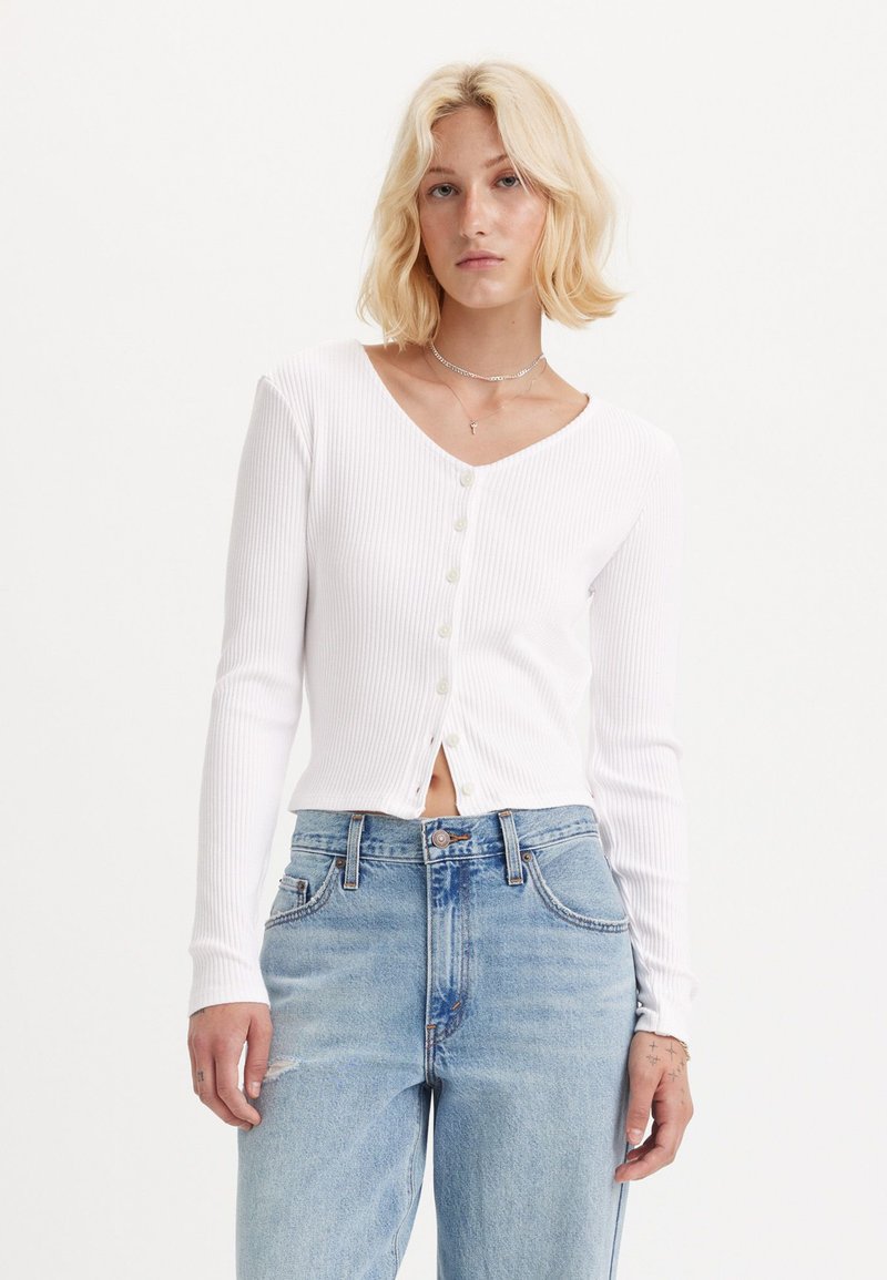 Top bianco a maniche lunghe con scollo a V e una pattina con cinque bottoni, abbinato a jeans azzurri a vita alta con dettagli strappati.