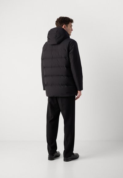 Veste rembourrée noire avec capuche, présentant des coutures horizontales et une coupe ample, assortie de pantalons et de chaussures noirs.