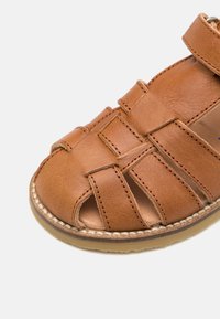 Sandalo in pelle color tan con un design a cinturino, aperto sia sulla punta che sul tallone. Accenti cuciti e una suola in gomma beige aggiungono dettagli.